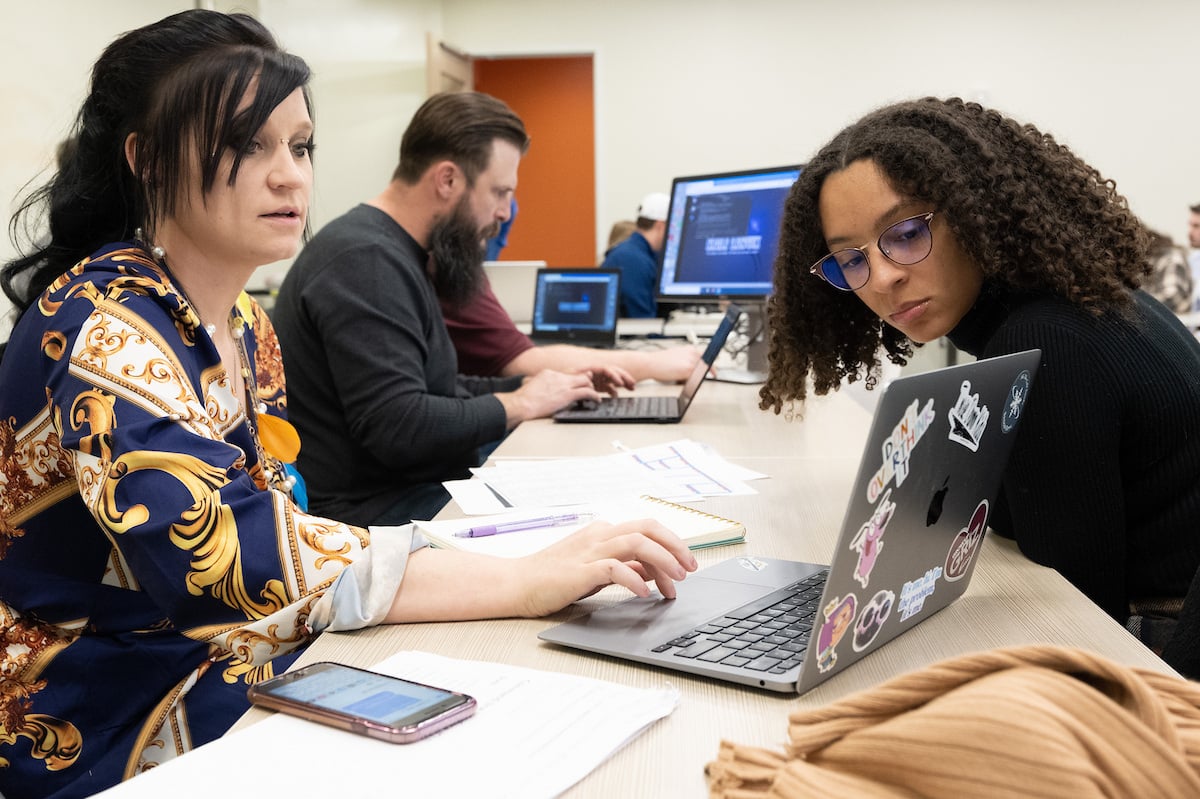 Two students sitting at a table across from each other studying on a laptop between them.