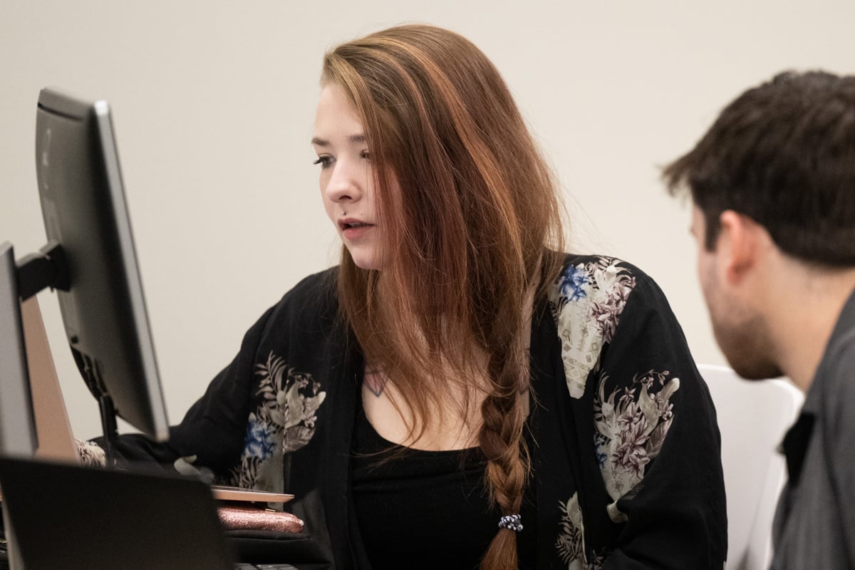 A UM student pointing at a computer monitor with another student looking on.