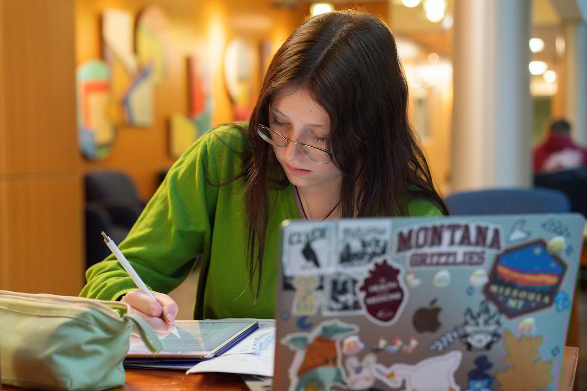 A UM student in a long sleeved green shirt sitting at a table taking notes next to a laptop