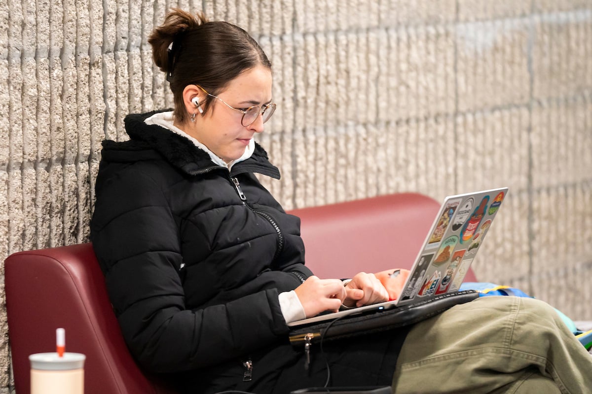 A UM student working on a laptop sitting on a maroon padded bench