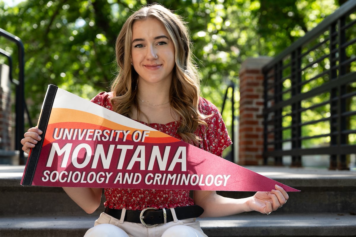 A UM student holding a University of Montana Sociology and Criminology pennant poses for a portrait on the University of Montana campus.