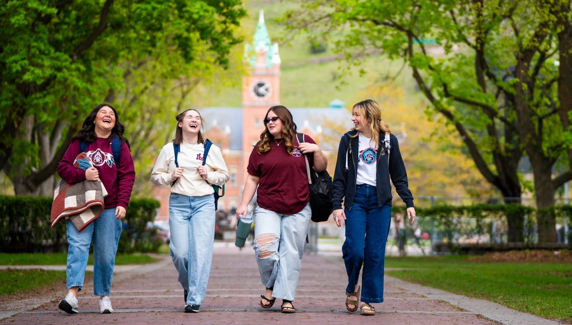 A group of four young women walks side by side on a picturesque brick path, sharing laughter and conversation with University of Montana's Main Hall in the background.